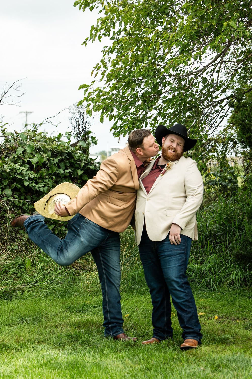 Two men in cowboy attire share a playful kiss on the cheek in a grassy outdoor setting, one lifting his leg and holding a straw hat.