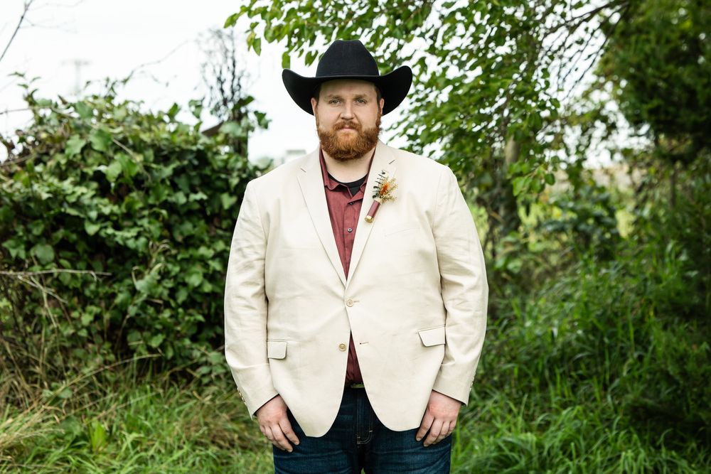 Bearded man in a black cowboy hat, cream blazer and boutonniere standing outdoors in front of green foliage.