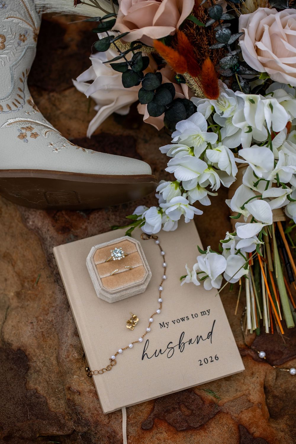 Wedding flat lay with a 2026 vows journal, ring box, pearl bracelet, white cowboy boot and a bridal bouquet of roses and white blooms.