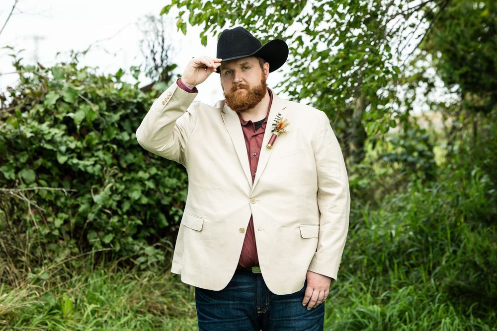 Bearded man in a black cowboy hat, cream blazer with a floral boutonniere and jeans stands outdoors tipping his hat before green foliage.