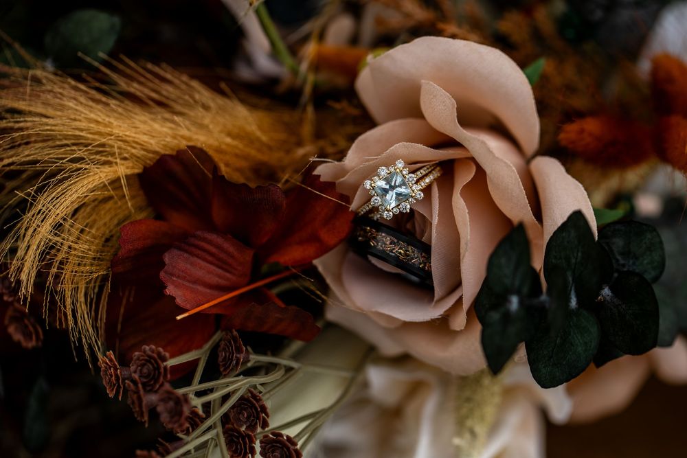Diamond engagement ring resting on a blush rose in a rustic autumn wedding bouquet with wheat stalks and dark burgundy flowers.