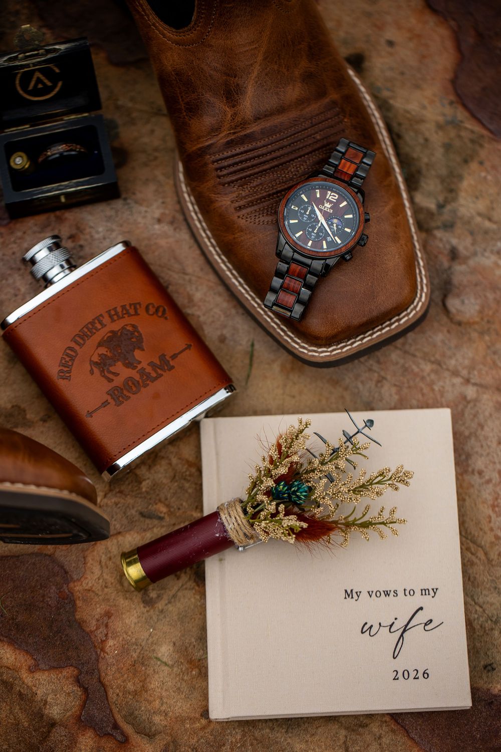 Groom's wedding flat lay with a leather flask, wood-band watch, cowboy boot, ring box, boutonniere and a vow book for 2026.