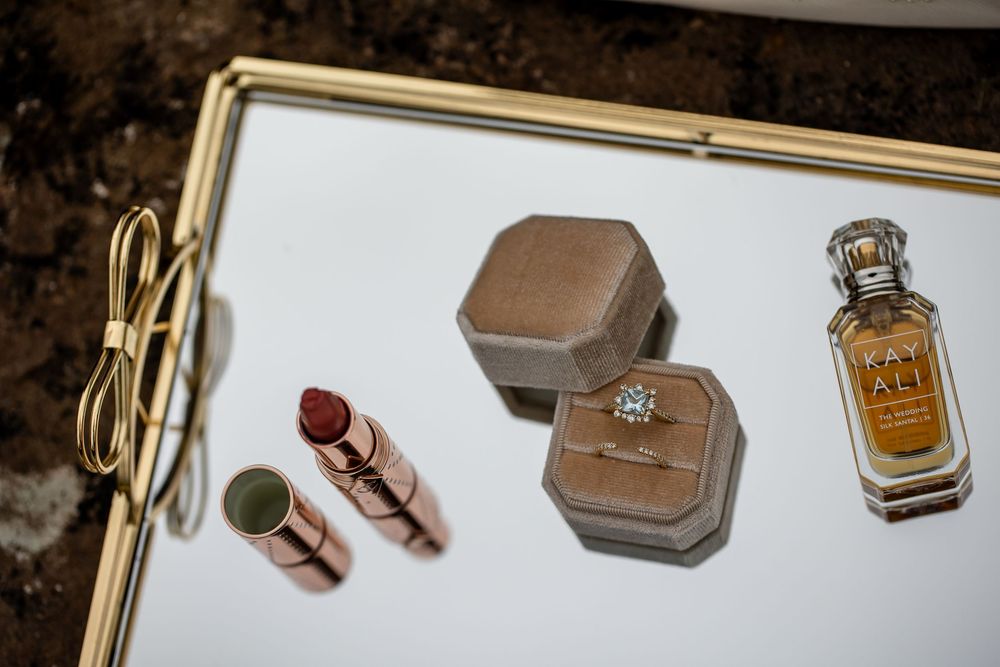 Diamond engagement ring in an open tan velvet box on a gold-framed mirrored tray beside lipsticks, a perfume bottle and a gold bow.