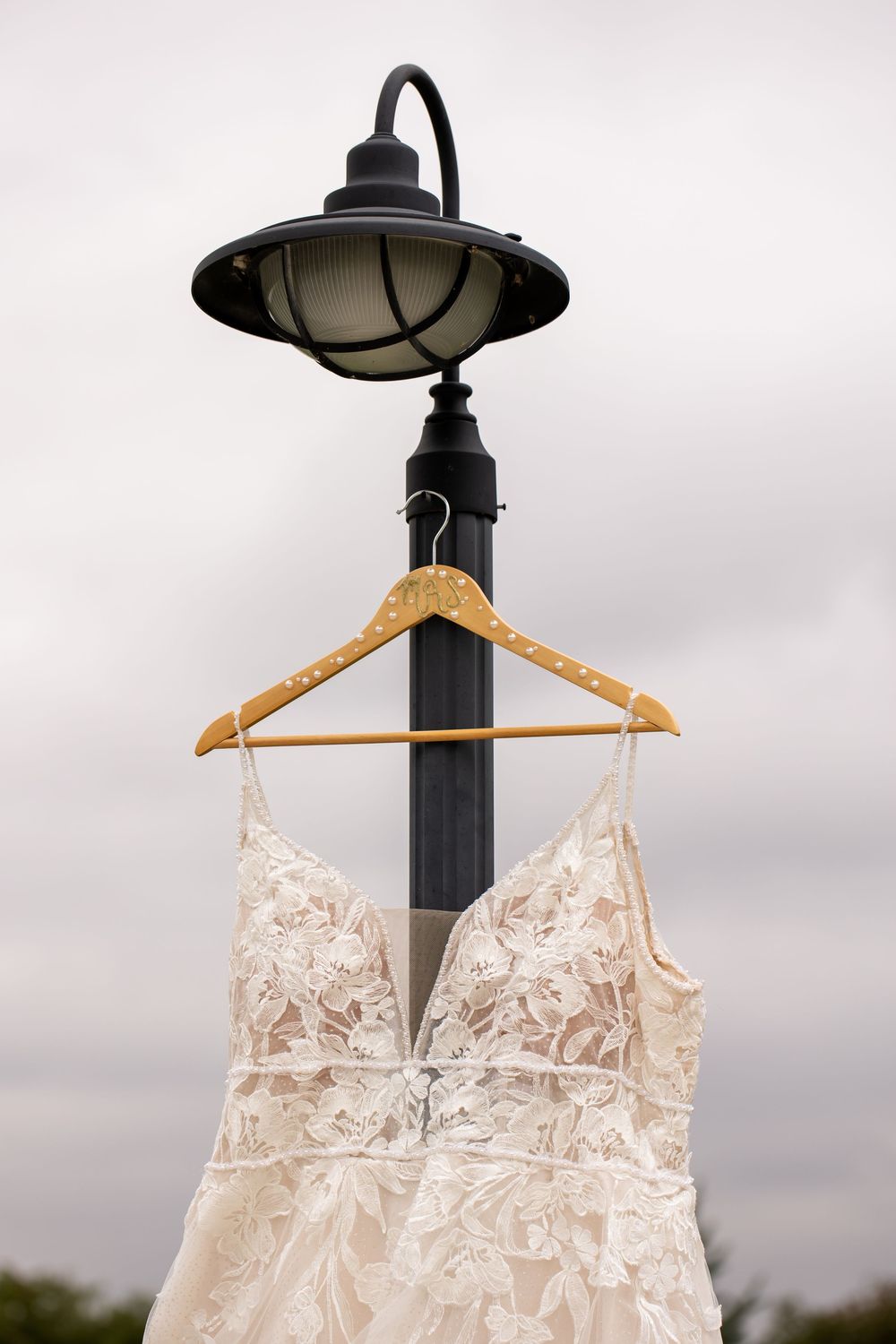 Ivory lace wedding dress with plunging neckline hanging on a wooden hanger from a black lamp post under a cloudy sky.
