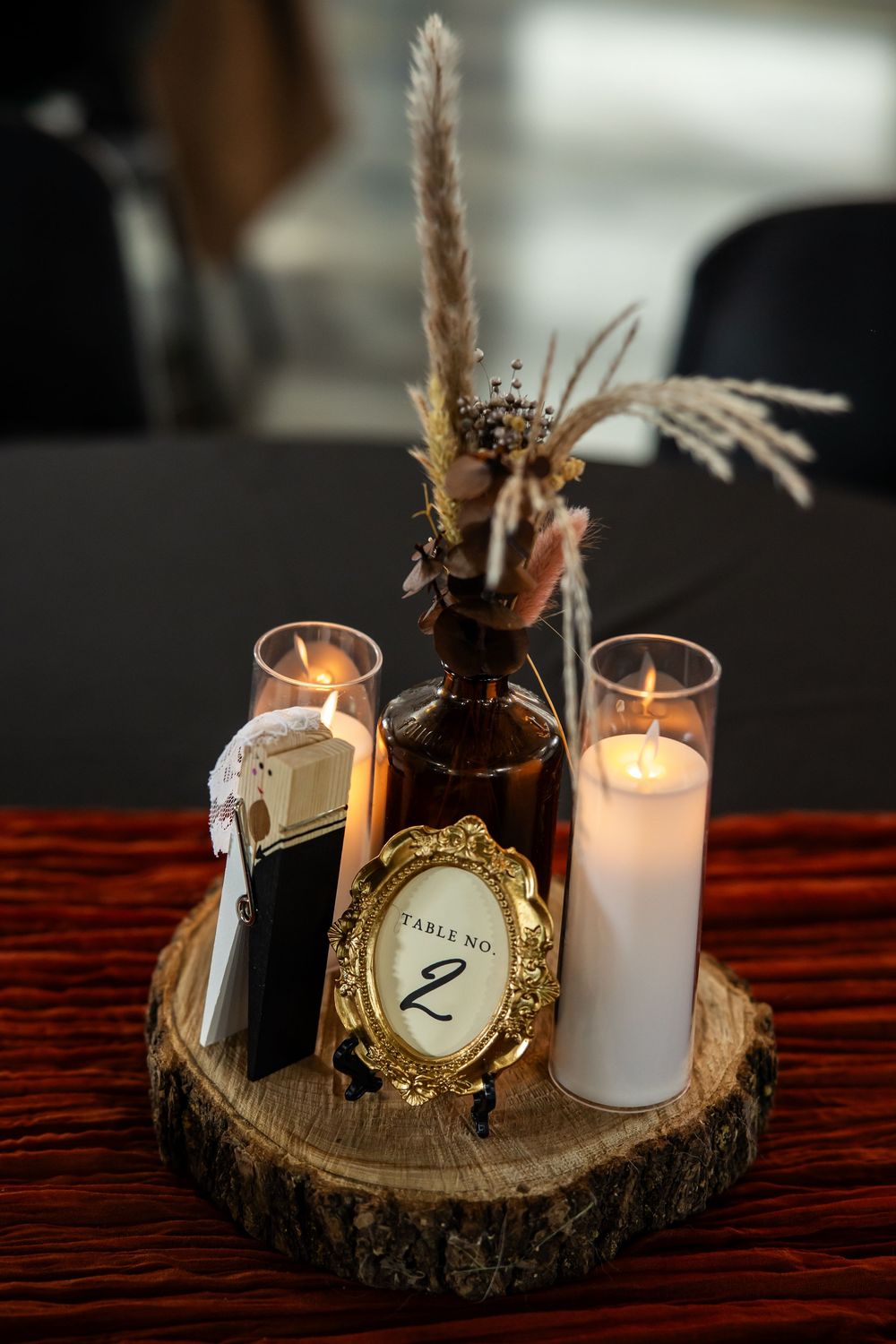 Wedding table centerpiece on a rustic wood slice with candles, pampas grass in a brown bottle, and a gold framed Table No. 2 sign.