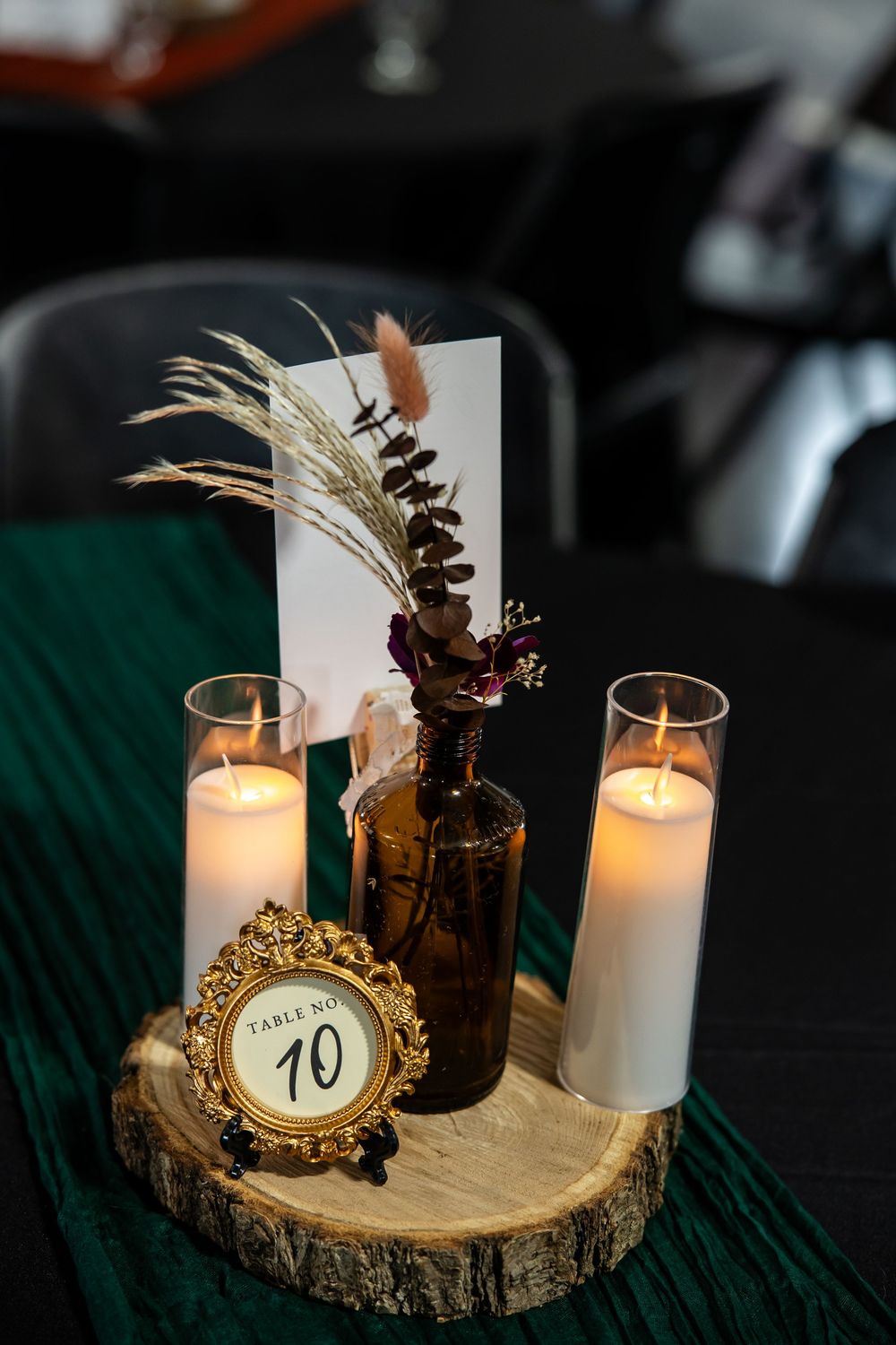Wedding table centerpiece with table number 10 in a gold frame, dried flowers in an amber bottle, and candles on a wood slice.