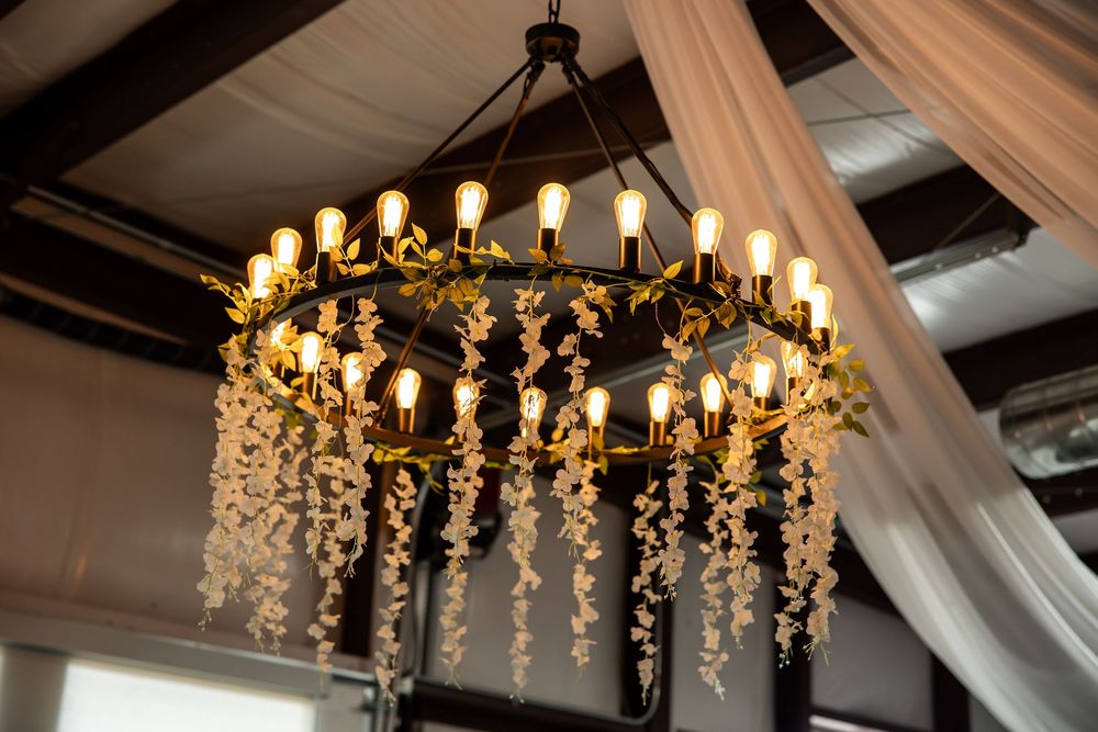 Round black wrought iron chandelier with Edison bulbs and cascading white floral garlands hanging beneath draped fabric at a wedding venue.