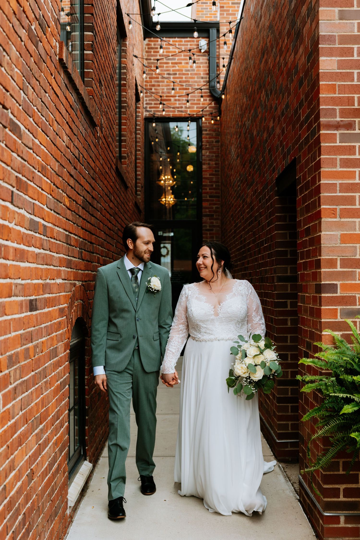 Bride and groom walk hand-in-hand through a narrow brick alley with string lights at their wedding.