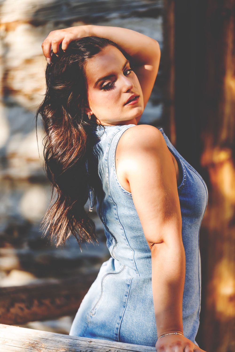 Young woman in denim dress poses outdoors, hand in dark hair, with rustic wooden background in warm sunlight.