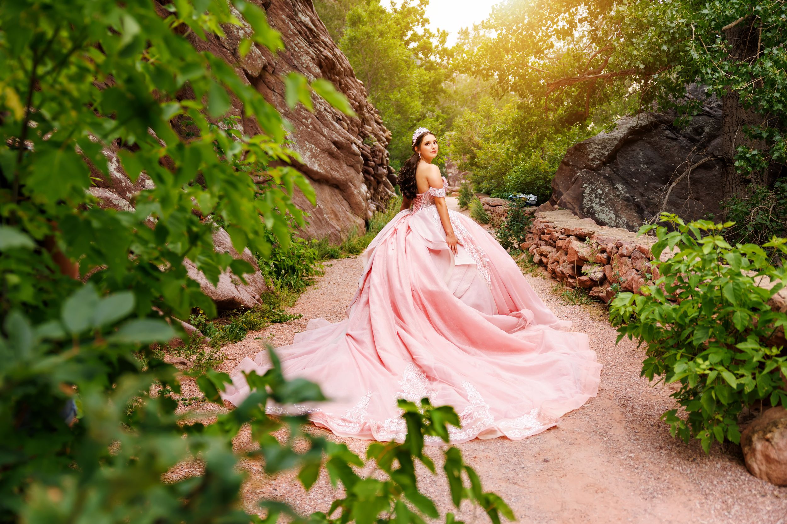 A woman in a flowing pink ball gown poses on a garden path surrounded by lush greenery and rocky cliffs.