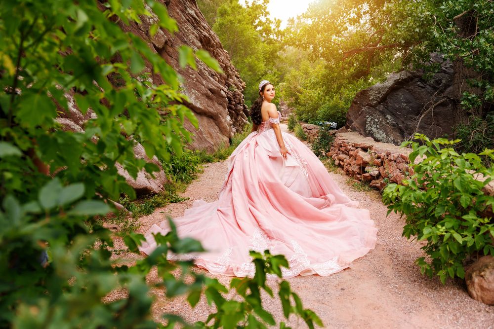 A woman in a flowing pink ball gown poses on a garden path surrounded by lush greenery and rocky cliffs.