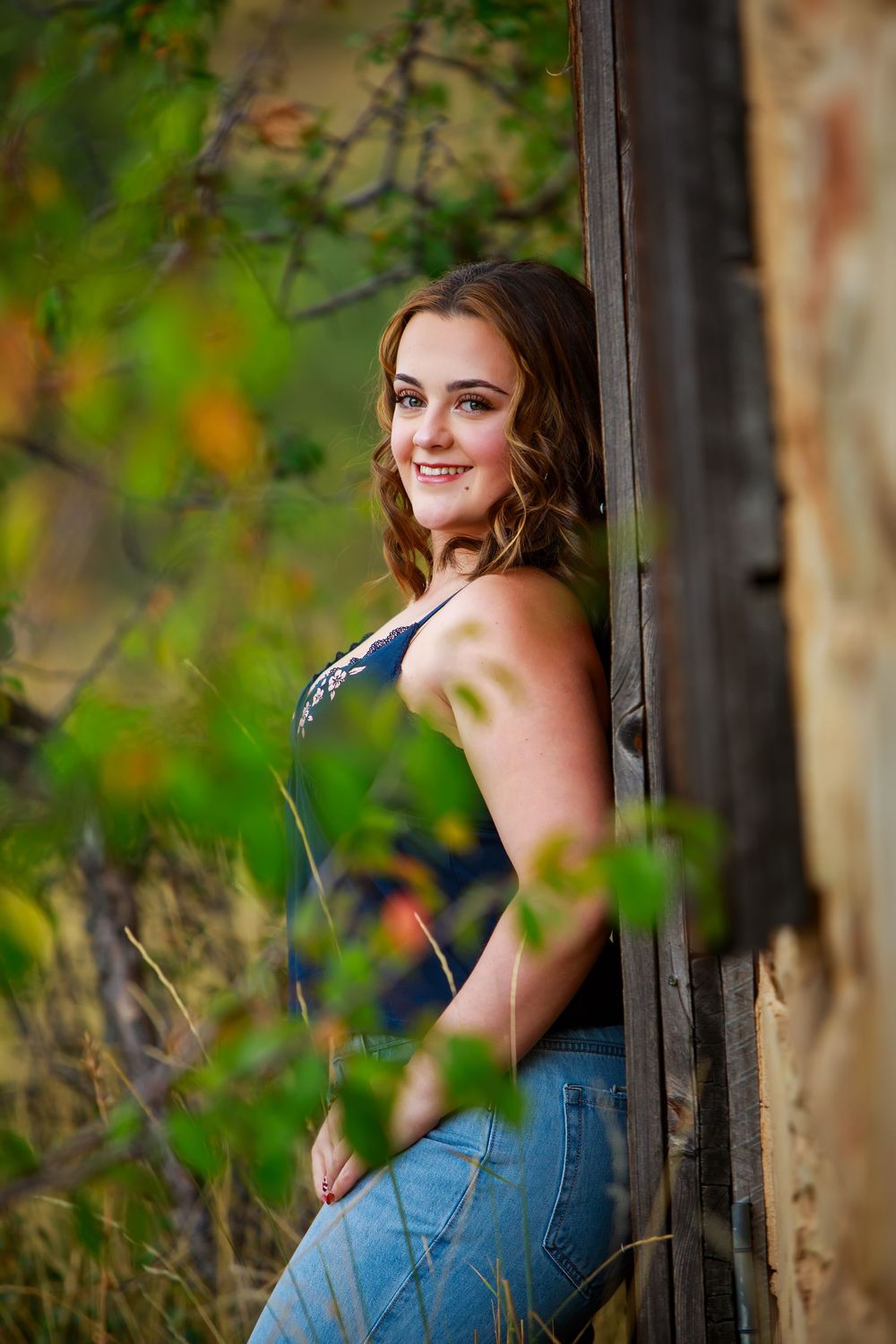 Young woman in jeans and tank top smiles while leaning against a rustic wooden structure surrounded by autumn foliage.