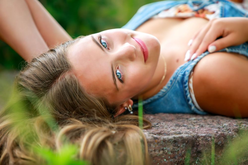 Young woman with blue eyes lying on a stone surface outdoors, smiling in a denim outfit surrounded by greenery.