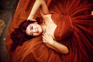 A figure in a flowing burgundy gown poses gracefully against a dark background in an artistic portrait series.