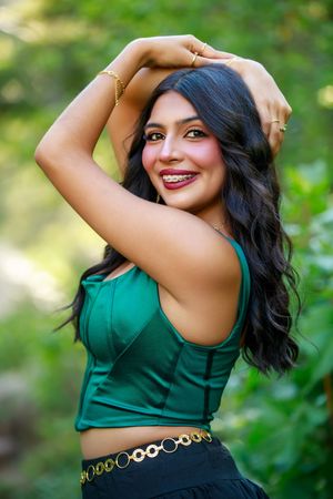 A person in a green sleeveless top and decorative belt poses outdoors against a blurred natural background.