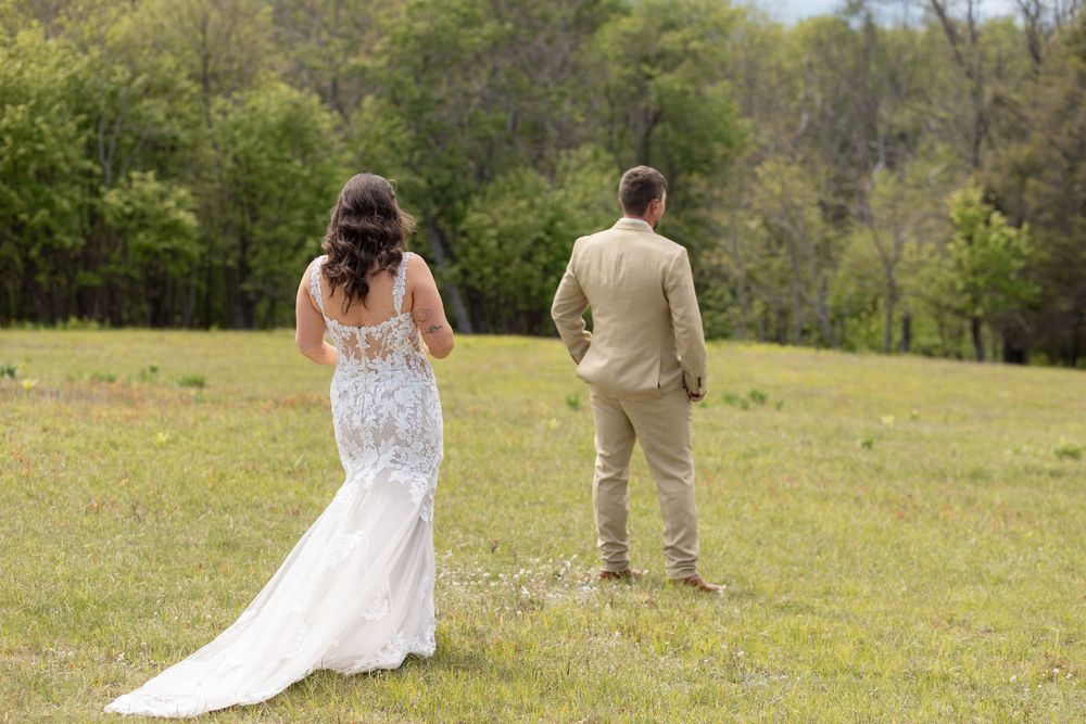 Bride in lace gown and groom in tan suit stand apart in green meadow during outdoor first look wedding photo.