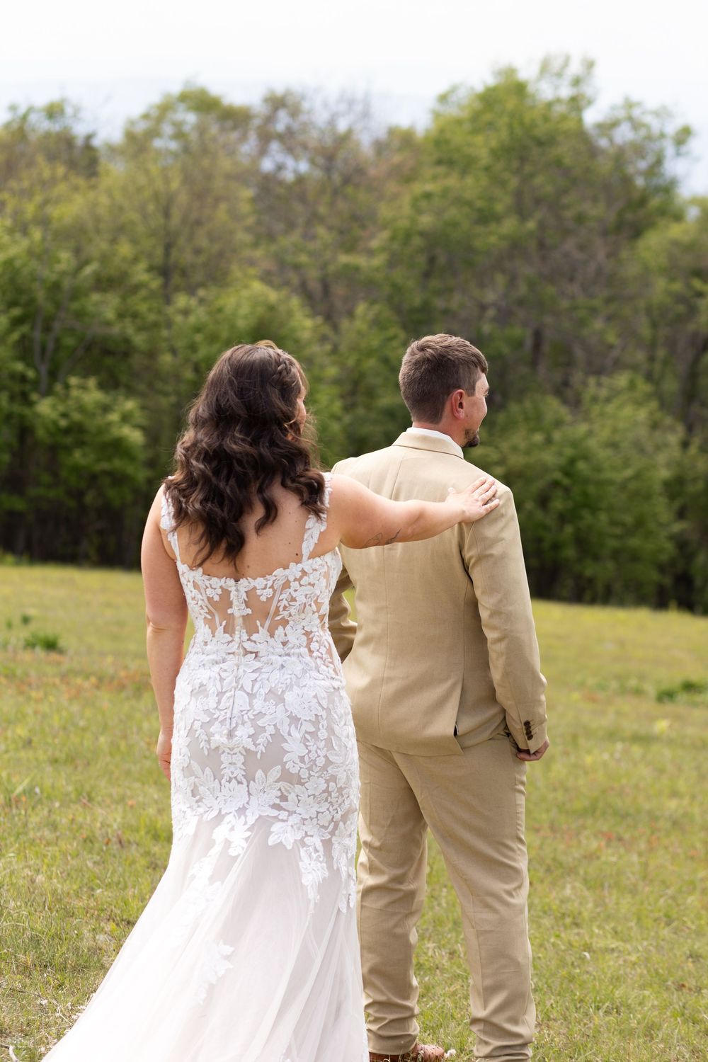 Bride in lace wedding gown and groom in tan suit stand together facing a lush green field and trees.