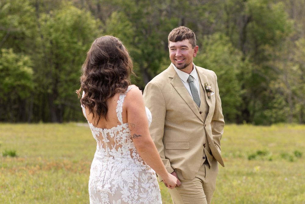 Bride and groom share first look moment in a lush green meadow, groom smiling in tan suit.