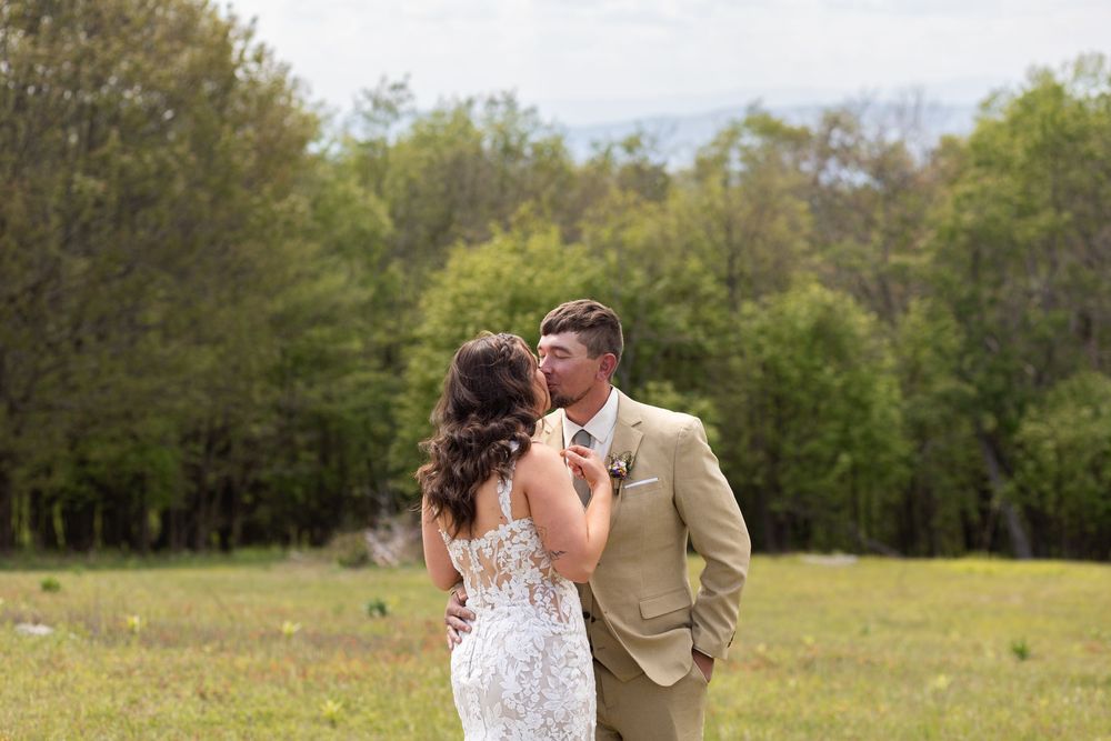 Bride and groom sharing a romantic kiss in a green meadow with lush trees and mountains in the background.