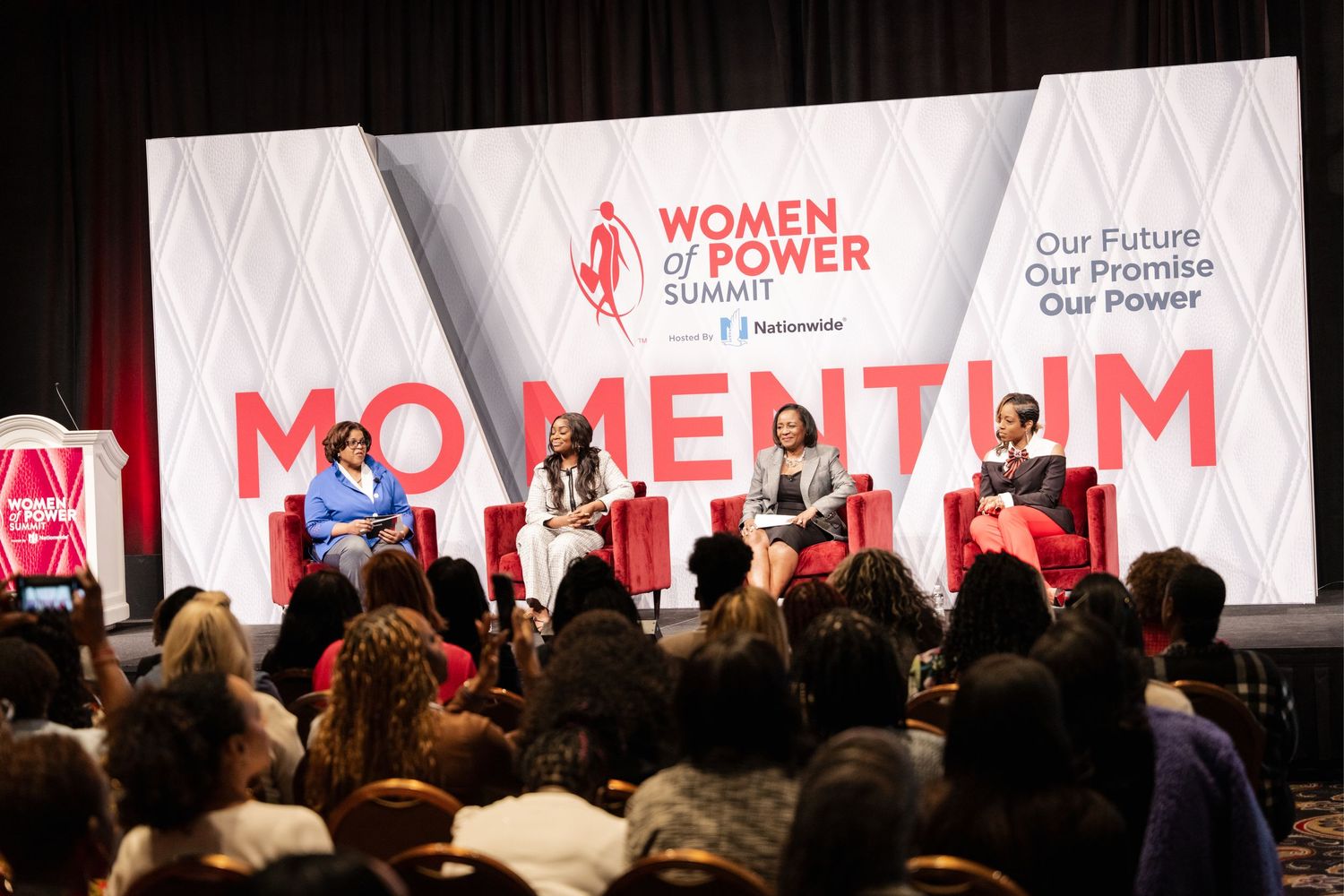 Two women speak on stage at the Women of Power Summit, sponsored by Nationwide, seated in red chairs.