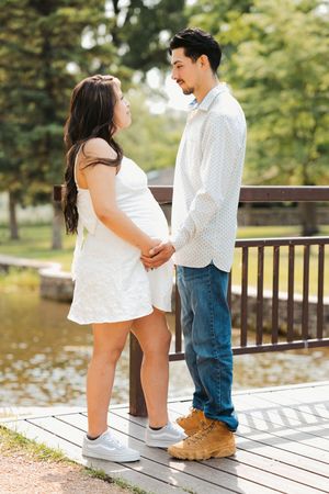 Pregnant couple holding hands on a wooden deck by a pond, woman in white dress, man in jeans and white shirt.