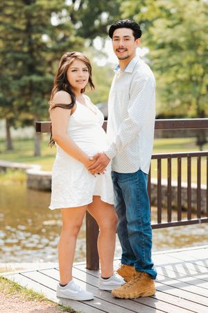A young couple holds hands on a wooden bridge in a park, the woman wearing a white dress and the man in jeans and a white shirt.