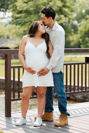 Pregnant couple posing on wooden bridge outdoors, man kissing woman's head, she wears white dress, both smiling softly.