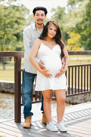 Couple posing on wooden bridge, pregnant woman in white dress, man in blue shirt and jeans, park setting.