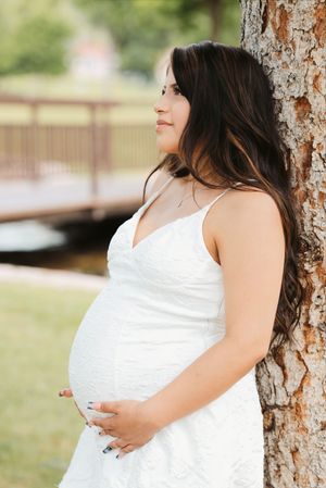 Pregnant woman in white lace dress leaning against tree in park, cradling baby bump, long dark hair, outdoor maternity photo.