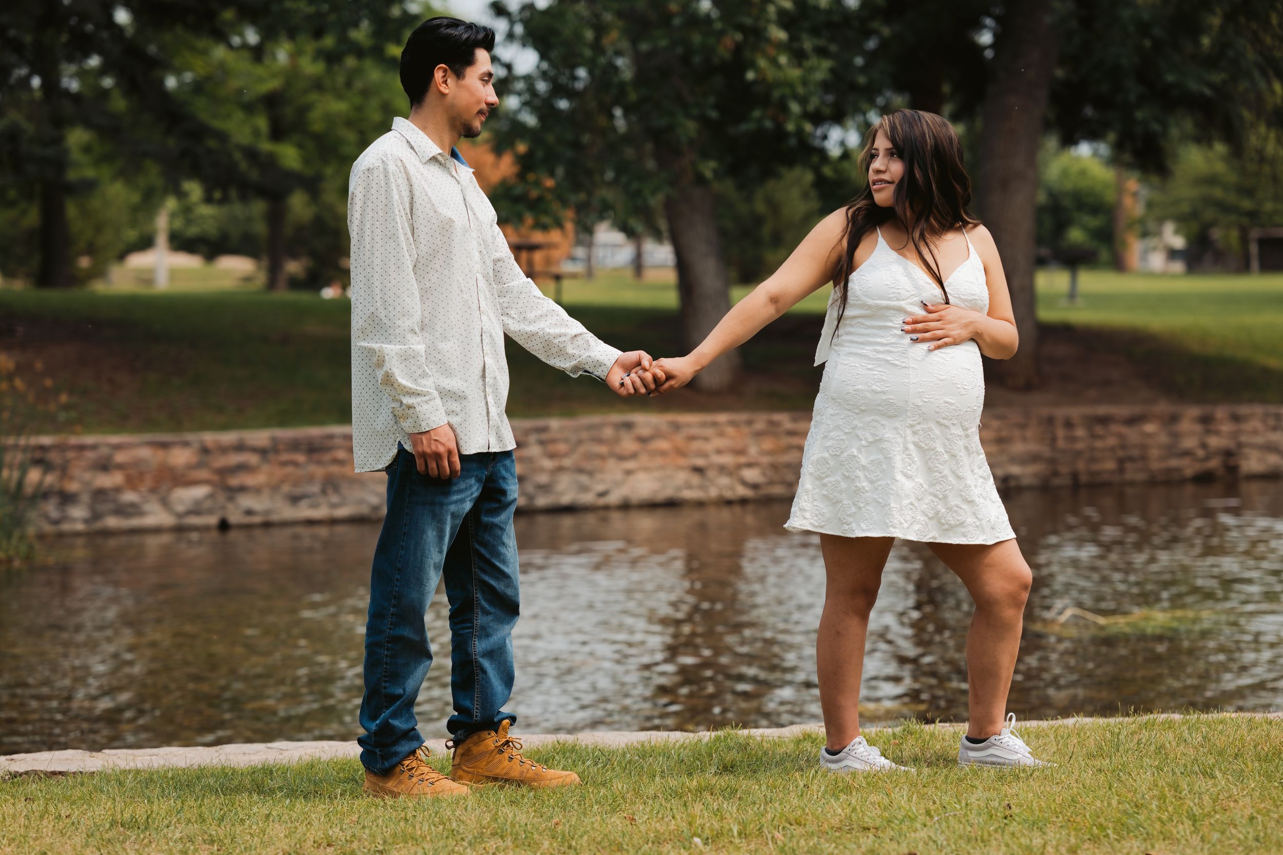 Pregnant woman in white dress holds hands with man near pond in park during maternity photo session.