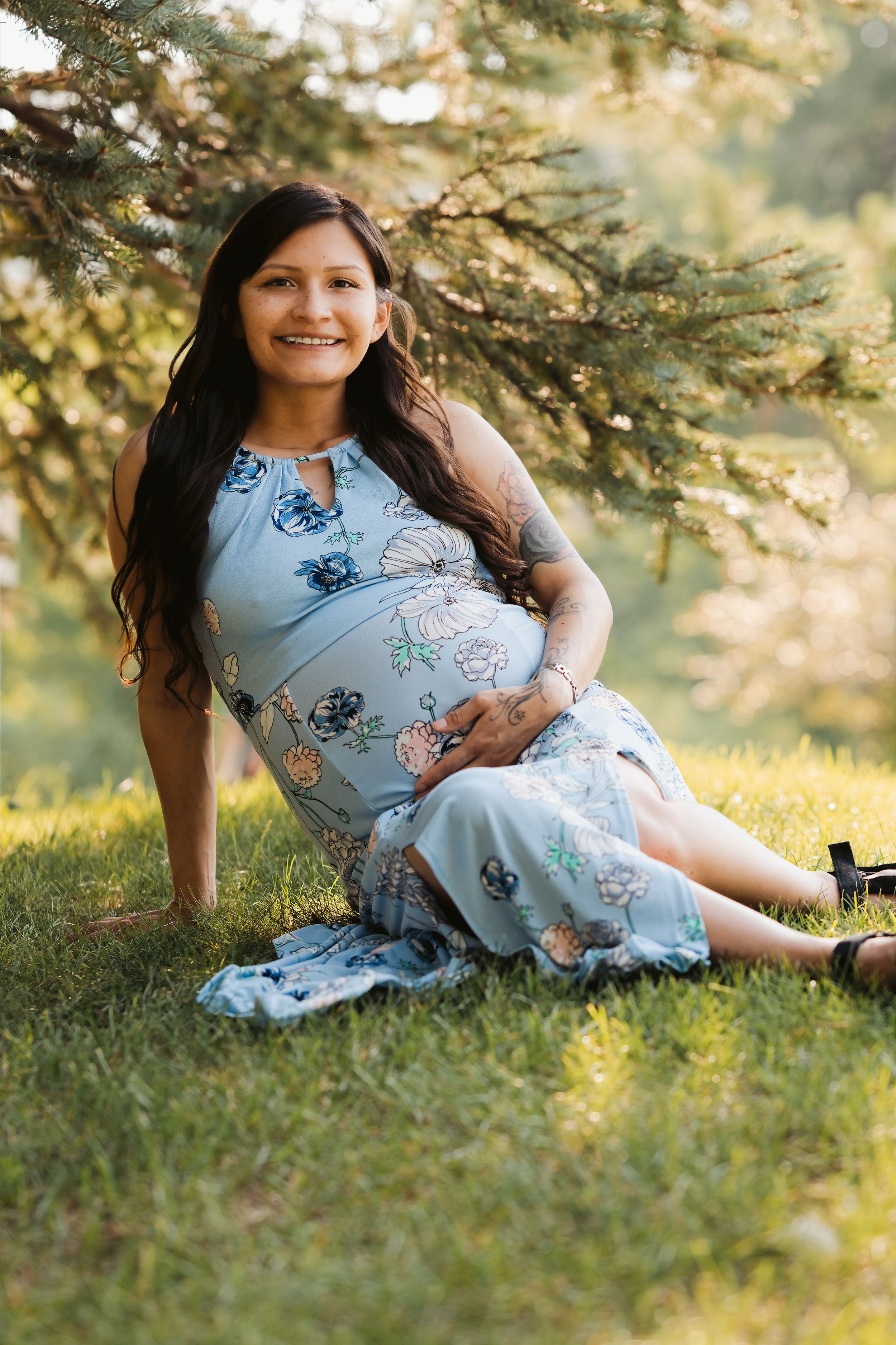Smiling pregnant woman in floral blue dress sitting on grass under pine tree in golden hour light.