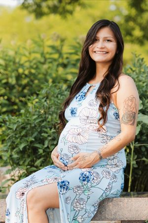 Smiling pregnant woman in floral blue dress sitting outdoors, cradling baby bump, with tattoos and green foliage background.