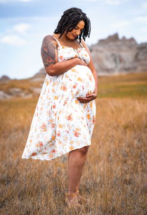 Pregnant woman in floral dress cradling baby bump in golden field with rocky badlands landscape backdrop.
