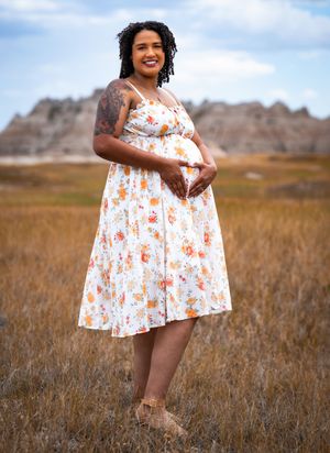 Pregnant woman in floral sundress poses in golden field with rocky Badlands formations in background.