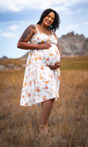 Smiling pregnant woman in floral dress holding baby bump in golden field with rocky formations behind her.