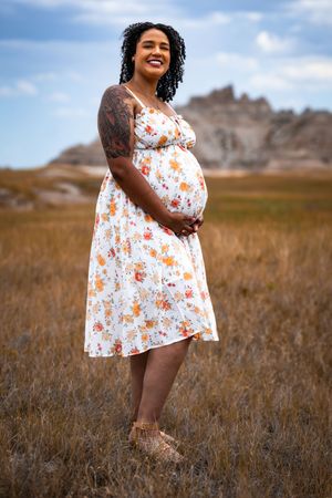 Smiling pregnant woman in floral dress standing in grassy field with rocky badlands formation in background.