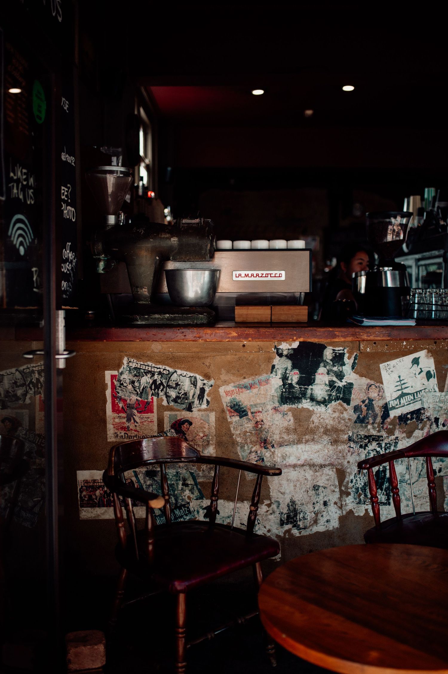 Dimly lit vintage cafe interior with worn posters, wooden chairs, and espresso machine on rustic counter.