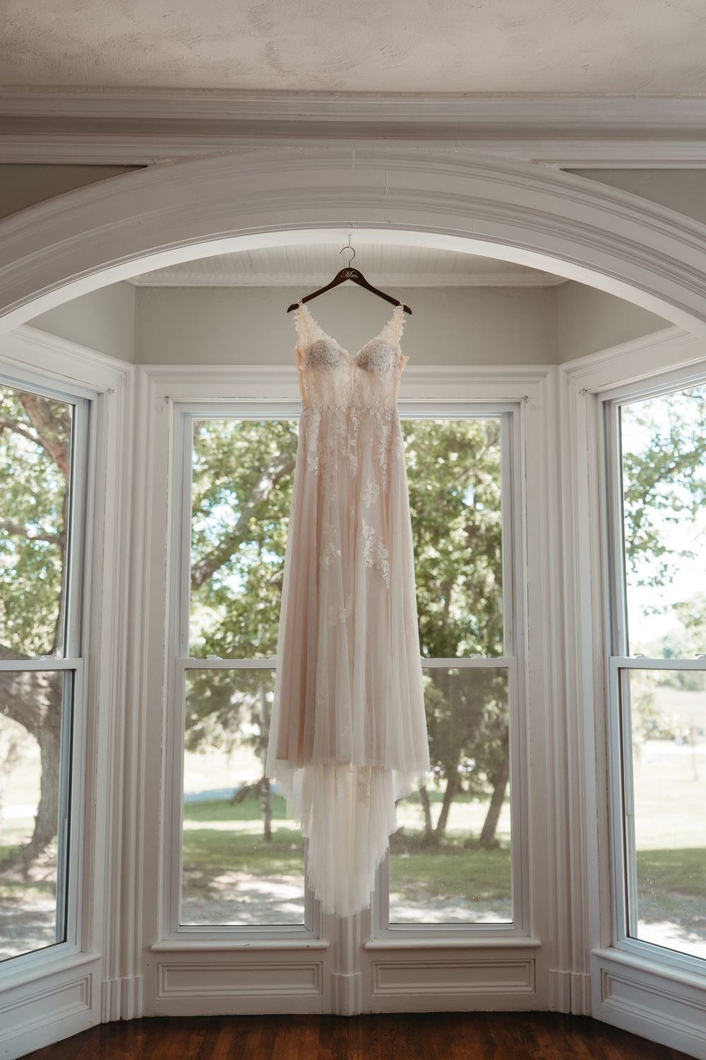 Elegant blush wedding dress with lace appliques hanging on a wooden hanger in a bay window.