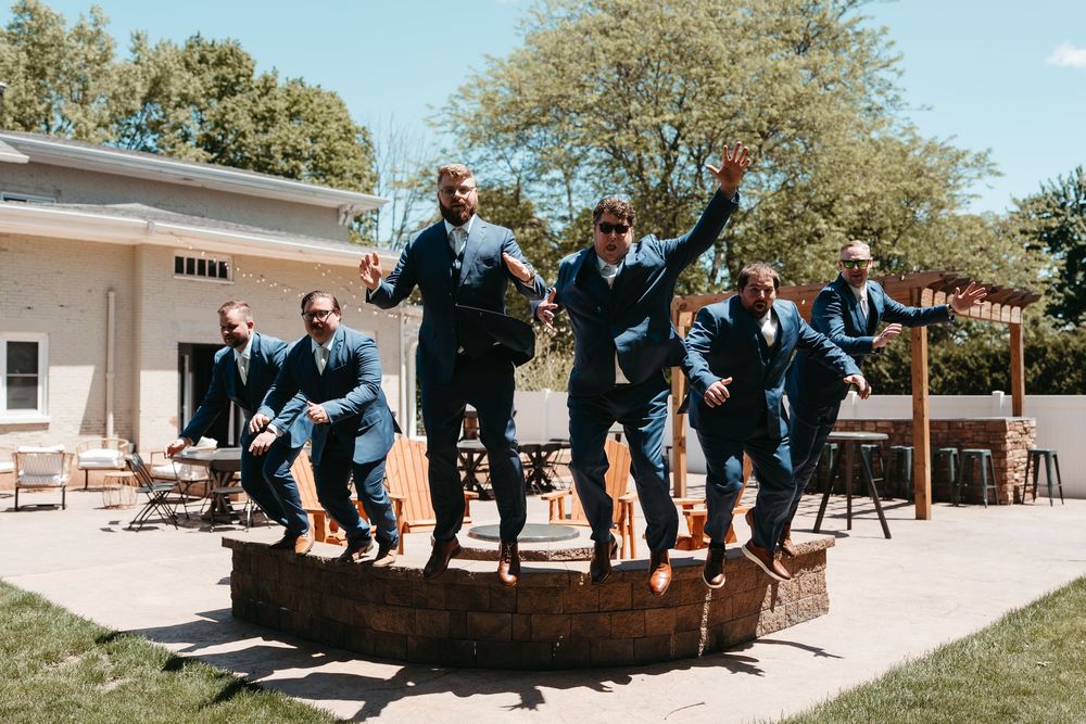 Groomsmen in navy suits jumping off a log outdoors at a wedding celebration.