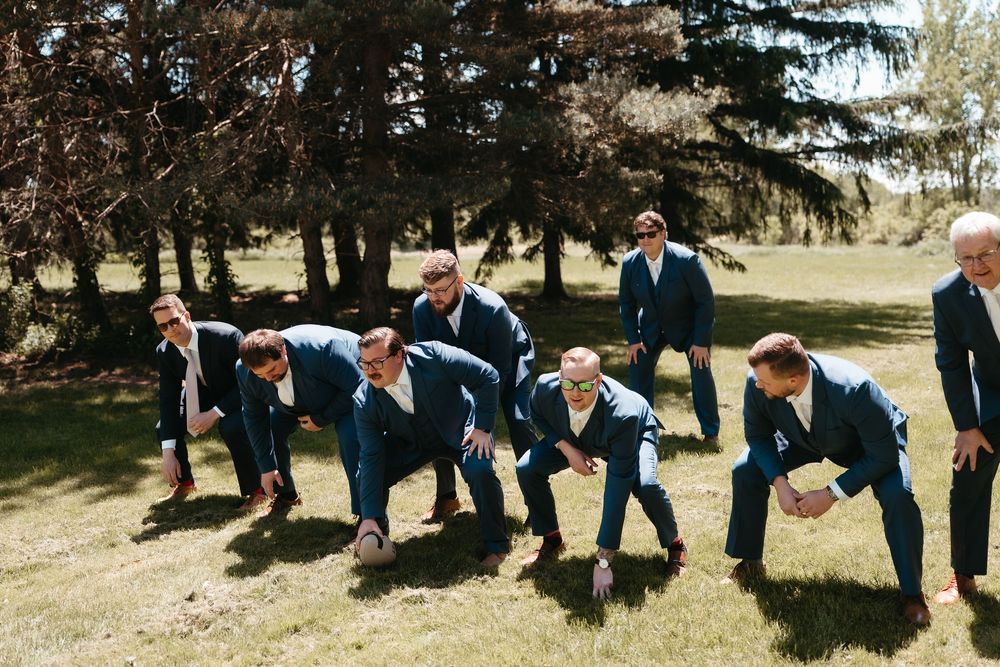 Groomsmen in navy suits pose playfully in a race stance on green grass at an outdoor wedding.