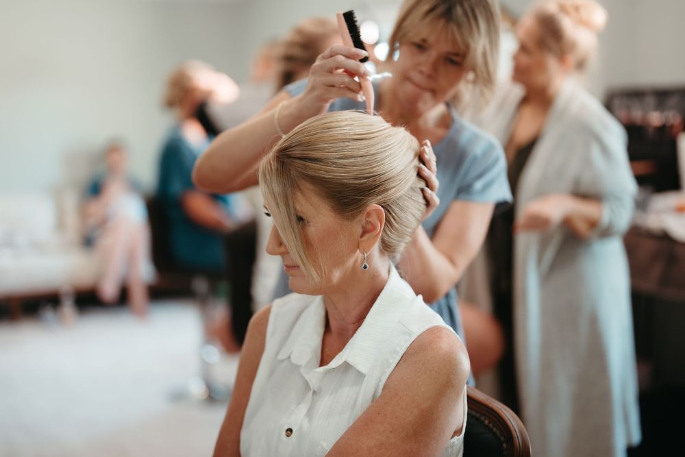 A hairstylist carefully styles an elegant updo on a seated woman during a bridal preparation session.