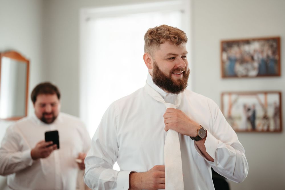 Groom in white dress shirt tying his tie while smiling, with groomsman on phone visible in mirror background.