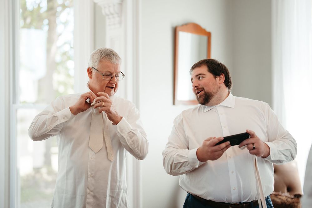 Two men in white dress shirts prepare for a wedding, one adjusting his tie while the other holds a phone.