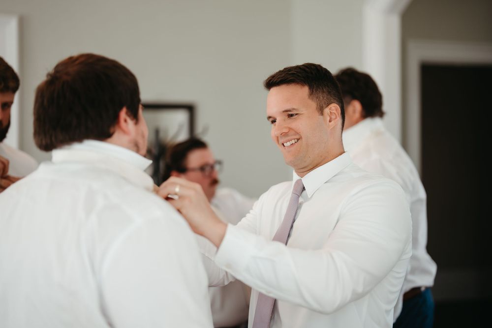Groomsmen in white dress shirts helping each other with boutonnieres while smiling during wedding preparations.