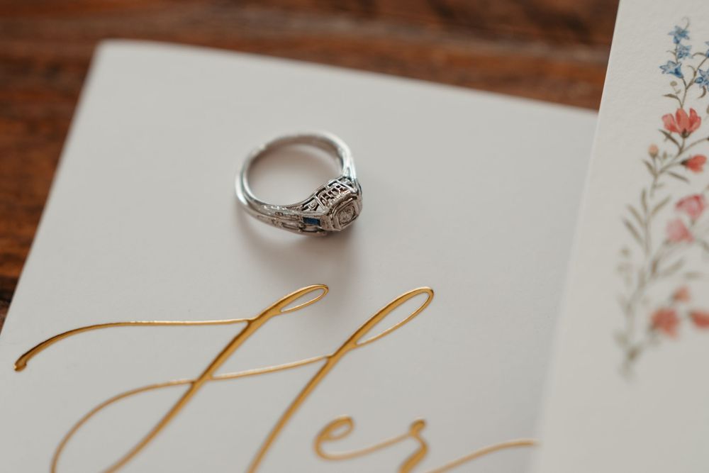 Elegant diamond engagement ring resting on a floral wedding card with gold script lettering.