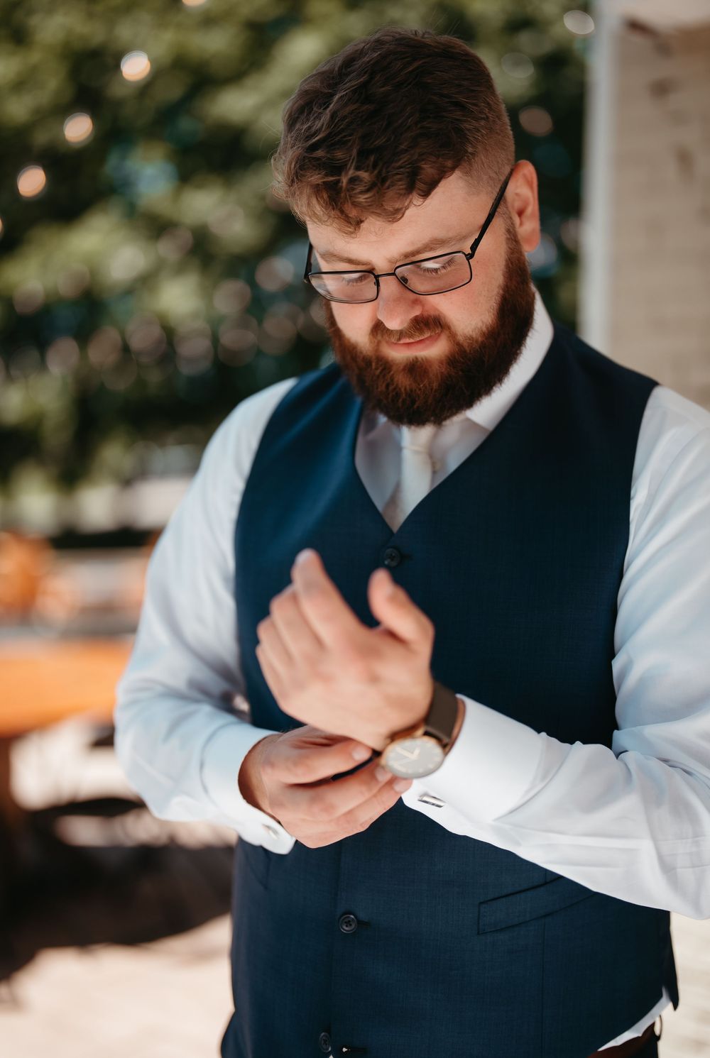 Bearded groom in navy vest and white shirt adjusting his cufflinks before the wedding ceremony.