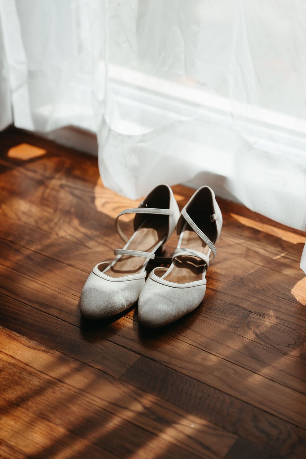 White bridal heels with cross-strap detail on wooden floor near sheer curtain in natural light.