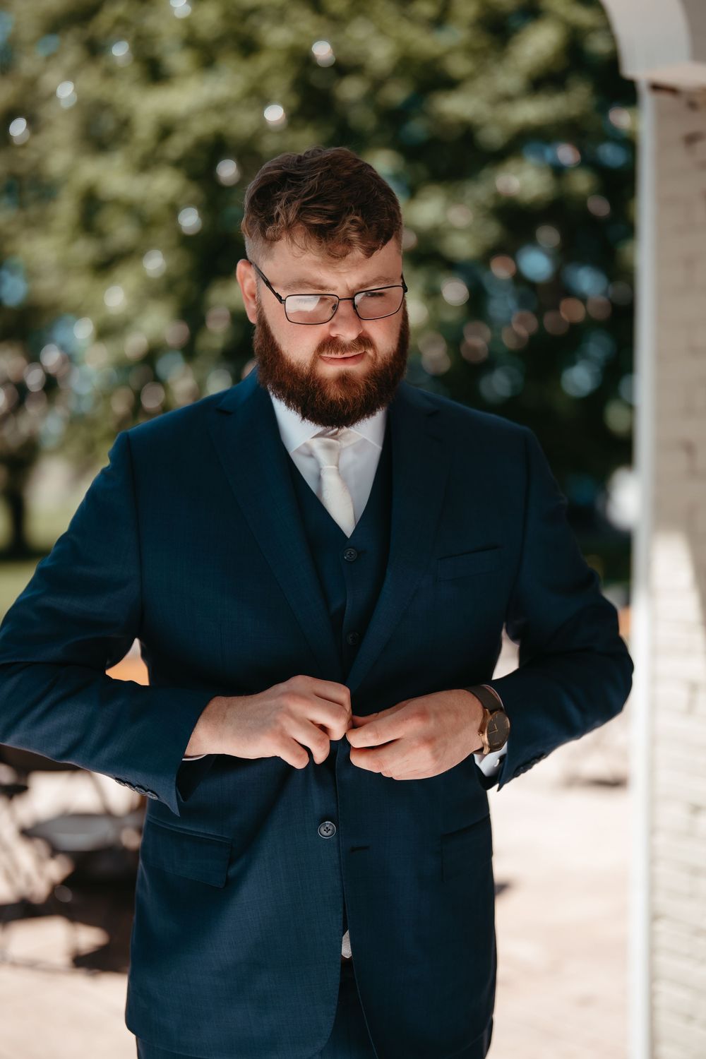 Bearded groom in navy three-piece suit adjusting jacket buttons outdoors on sunny wedding day.