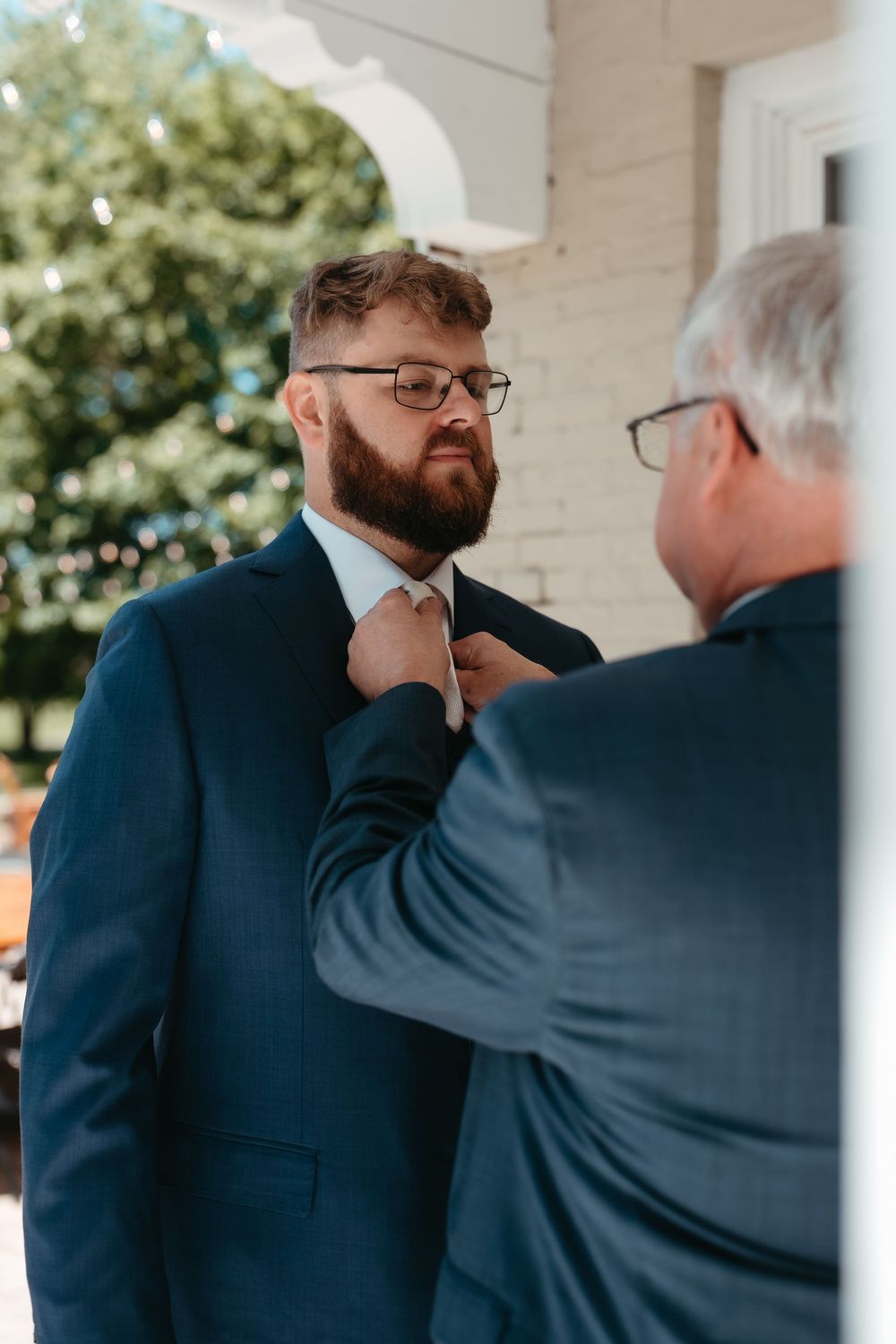 A bearded groom in a navy suit gets his tie adjusted by an older man in glasses outdoors before a wedding ceremony.
