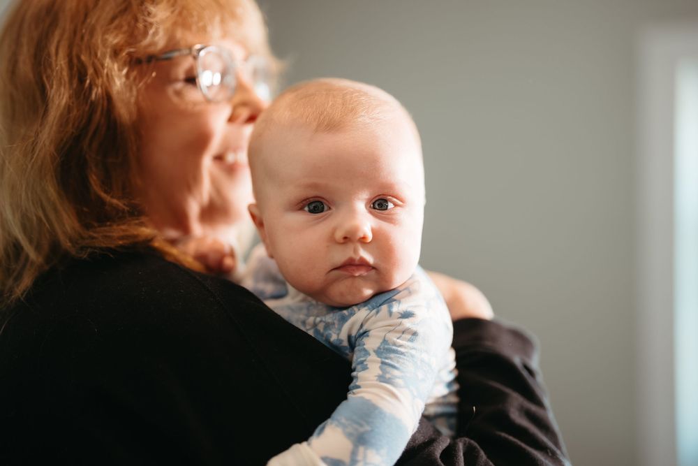 Grandmother with glasses lovingly holds a wide-eyed baby in a blue outfit, sharing a tender moment together.